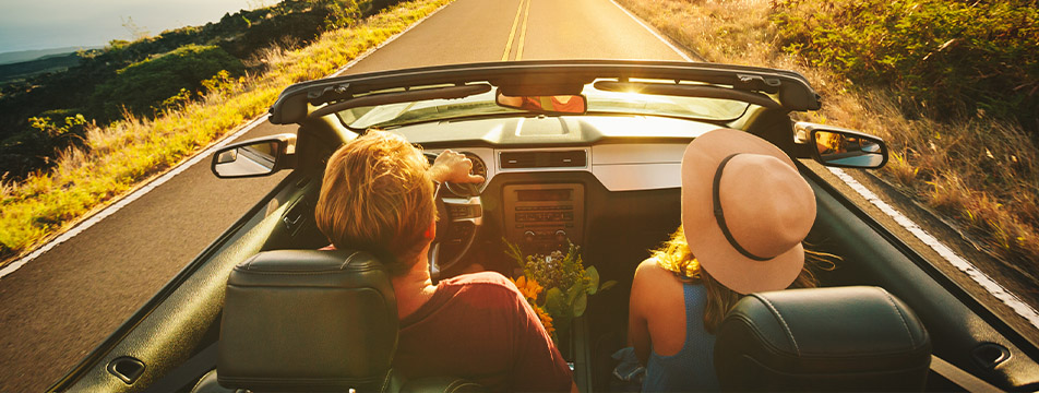 Couple driving a convertible car on a sunny open road surrounded by nature, with the woman wearing a hat and holding flowers.