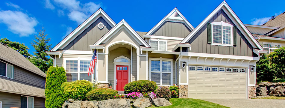 Front view of a modern suburban house with gray siding, a red front door, an American flag, a two-car garage, and well-maintained landscaping under a clear blue sky.
