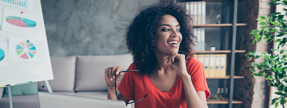 Woman in a red shirt holding eyeglasses and sitting at a desk with a laptop, with a chart on a whiteboard and shelves in the background.