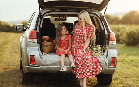 Woman and young girl sitting on the open tailgate of a silver SUV in a grassy field during sunset, both wearing red polka dot dresses, with a basket and flowers beside them.