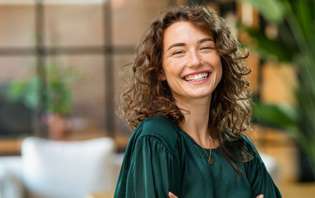 Woman with curly hair wearing a green blouse sitting indoors with arms crossed, with a blurred background of a modern living space.