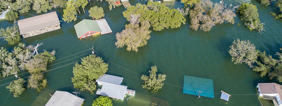 Aerial view of a flooded residential area with houses partially submerged in water and trees scattered throughout.