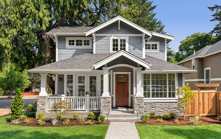 Two-story gray house with white trim, a front porch with white railings, and a well-maintained lawn and garden under a clear blue sky