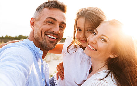 Smiling family selfie of a father, mother, and young daughter outdoors at sunset — the father holds the camera while all three lean close together and look at the lens.